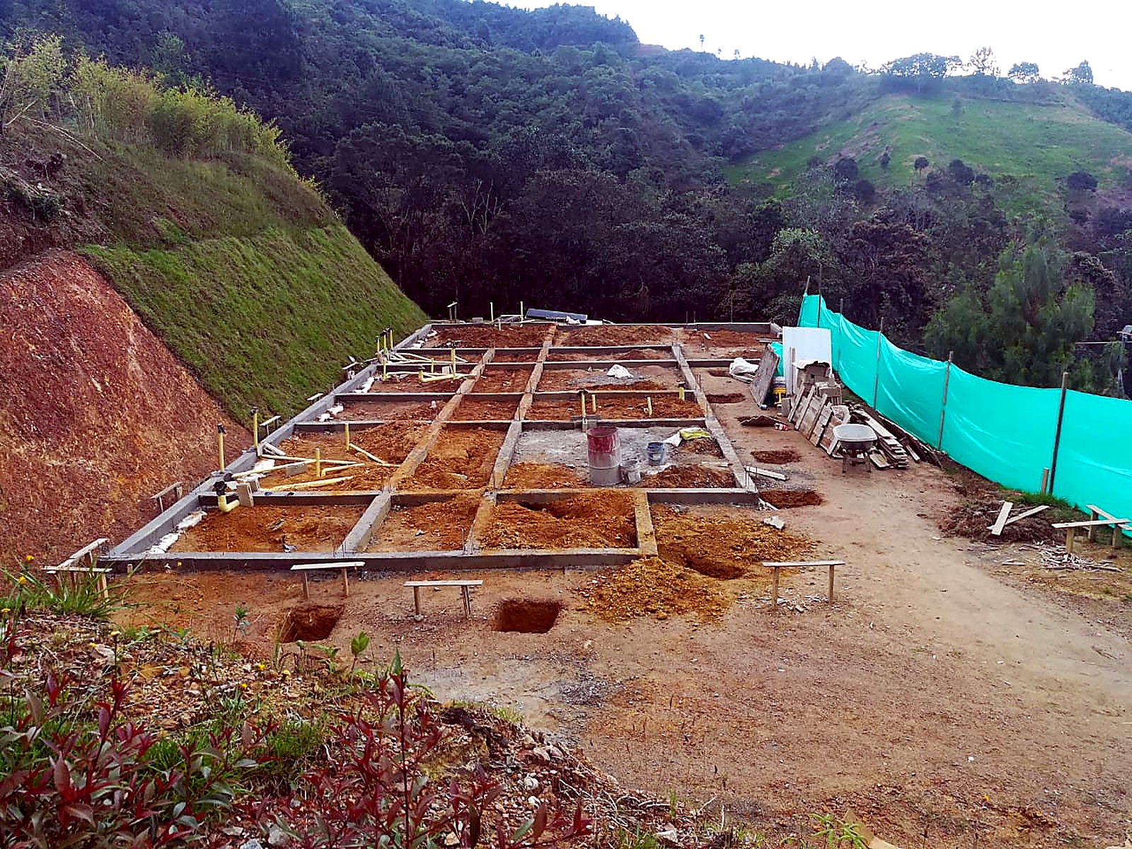 Vigas de cimentación fundidas y excavaciones de zapatas frente al cerramiento verde
